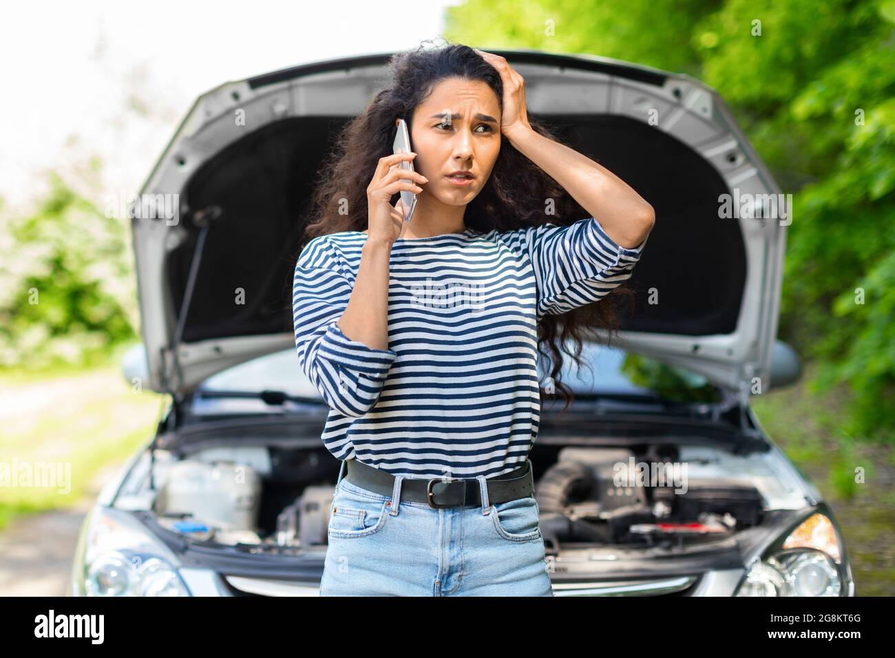 Nervöse Frau mit offener Autohaube, die den Autoservice anruft Stockfoto
