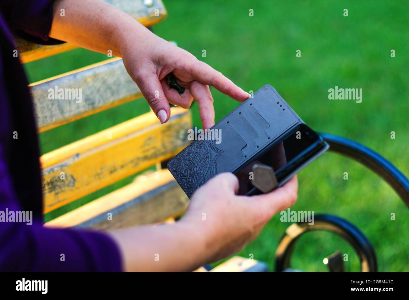 Unschärfe-Unschärfe-Nahaufnahme weibliche Hände suchen auf dem Telefon außerhalb, im Freien. Frau in lila Bluse mit Smartphone, während sie auf einer Bank im Park sitzt. C Stockfoto
