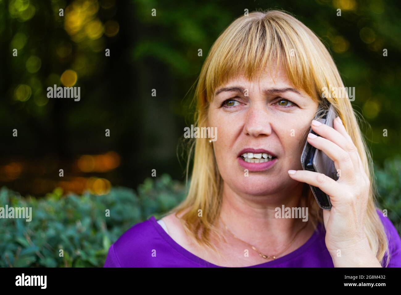 Unschärfe-Nahaufnahme kaukasisch blonde Frau sprechen, sprechen auf dem Telefon draußen, im Freien. 40-jährige Frau in lila Bluse im Park. Erwachsene Frauen usin Stockfoto