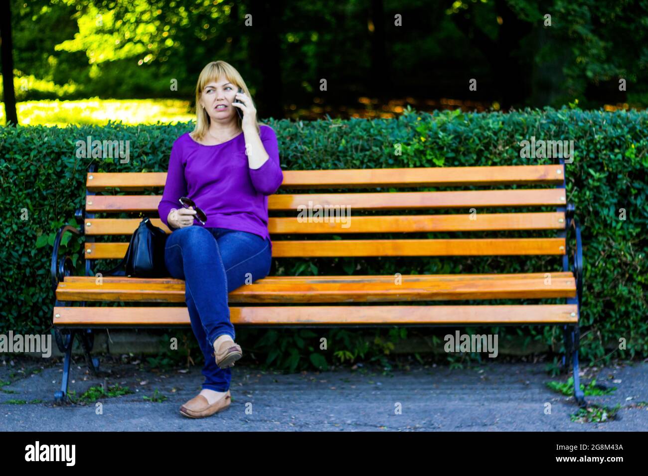 Unschärfe kaukasisch blonde Frau reden, sprechen am Telefon draußen, im Freien. 40-jährige Frau in lila Bluse im Park auf der Bank. Erwachsene Frauen usin Stockfoto