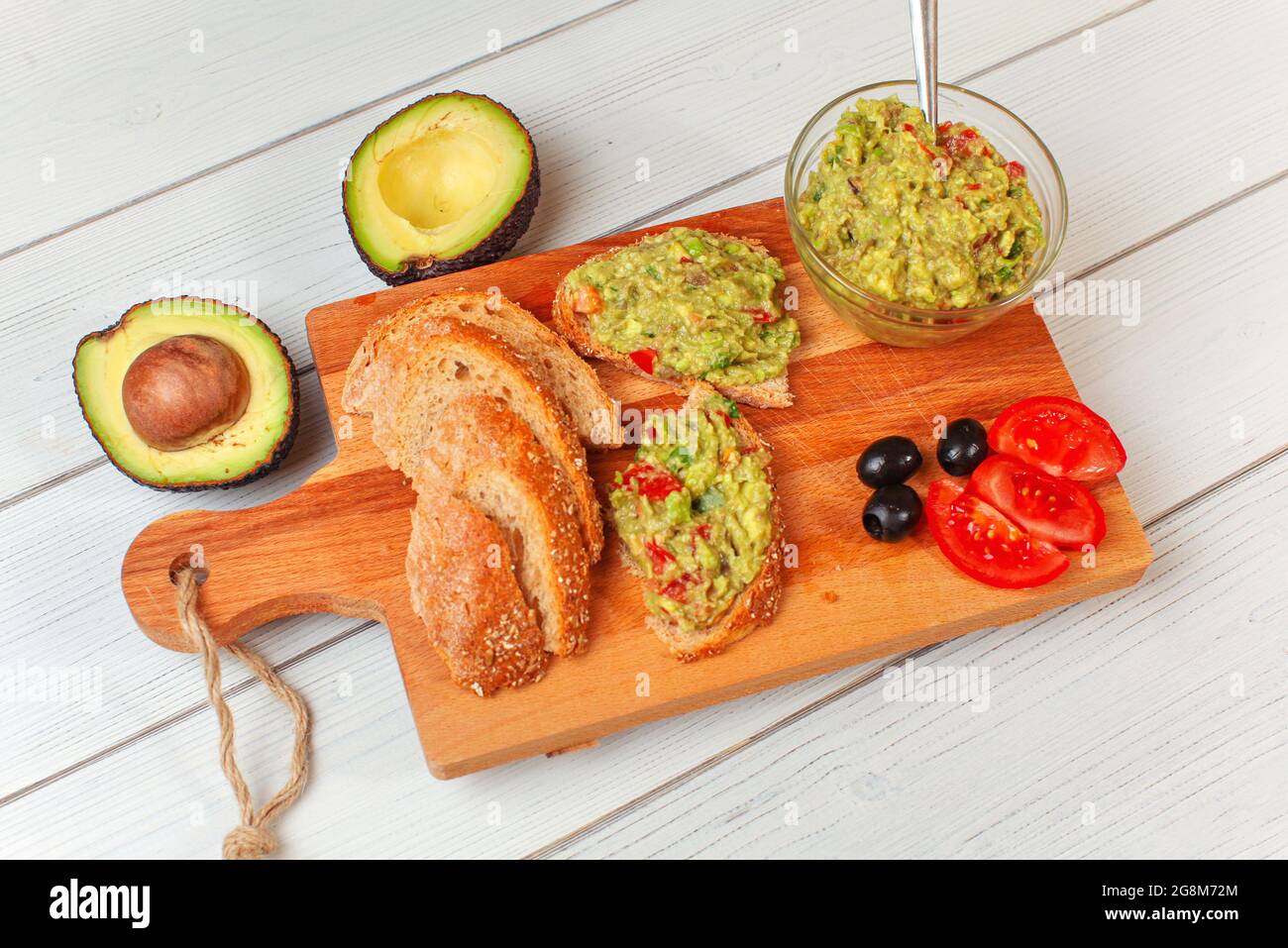 Frisch zubereitete Guacamole in kleiner Glasschüssel, Brot, Tomaten, Oliven am Arbeitsbrett und zwei Avocados-Hälften weiter, Blick von oben Stockfoto