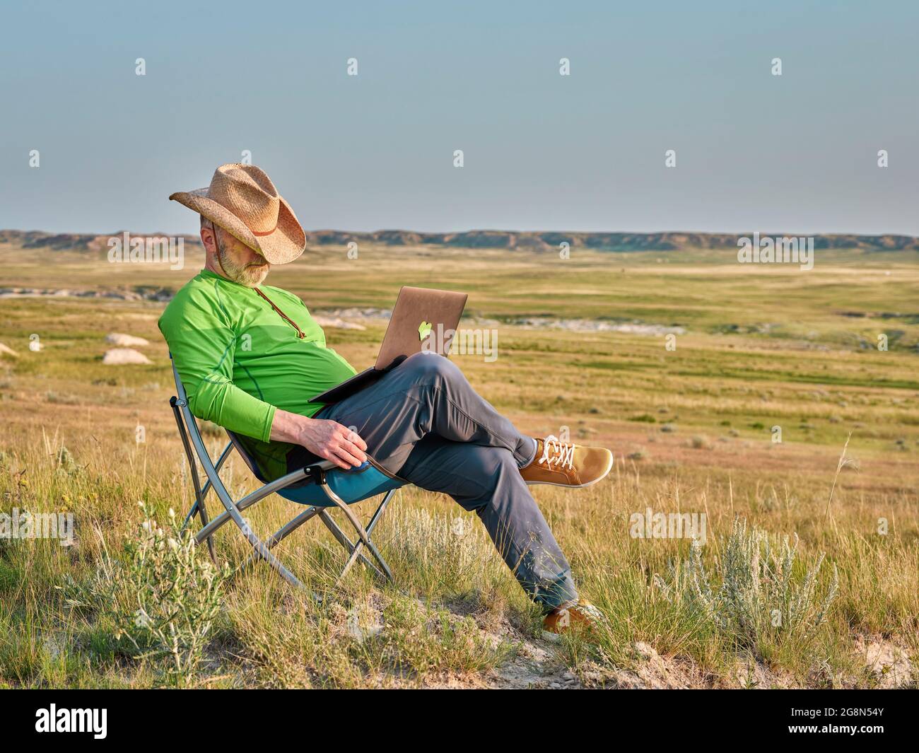 Ein älterer Mann mit Cowboyhut macht ein Nickerchen in einem Klappstuhl, während er mitten im Nirgendwo, am frühen Morgen in Pawnee National Grassla, am Laptop arbeitet Stockfoto