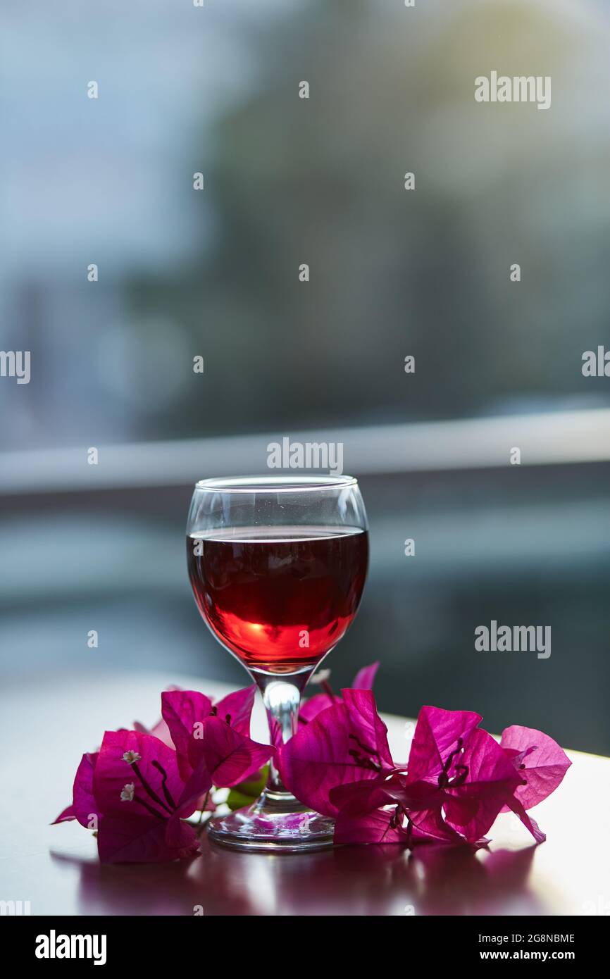 Ein Glas Rotwein auf unscharfem Berghintergrund. Dekorationen von rosa Bougainvillea Blumen. Blue Hour Fotografie. Speicherplatz kopieren. Stockfoto