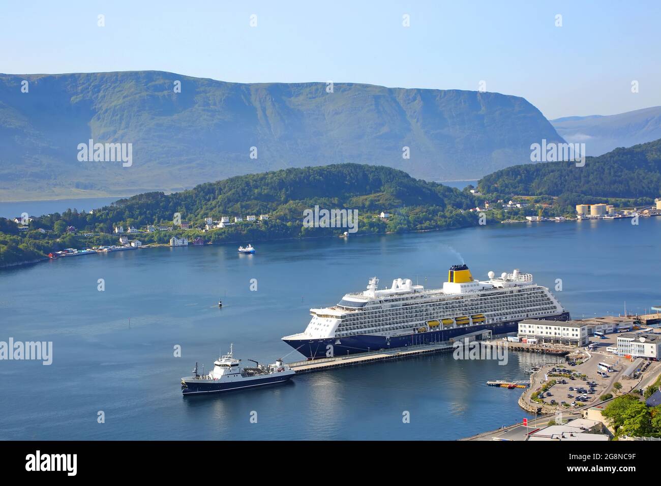 Das Kreuzschiff mit blauem Rumpf und gelbem Trichter dockte im Hafen von Alesund, Norwegen, an. Schöne Fjordlandschaft mit Berg im Hintergrund Stockfoto