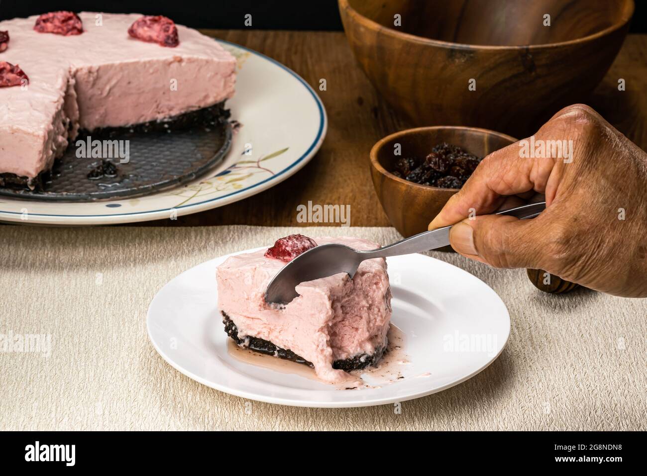 Senior Hand mit Metalllöffel schneiden köstliche gefrorene hausgemachte Erdbeere Käsekuchen in weißen Keramikschale auf Tischmatte mit getrockneten Rosinen in Holzbecher, Stockfoto