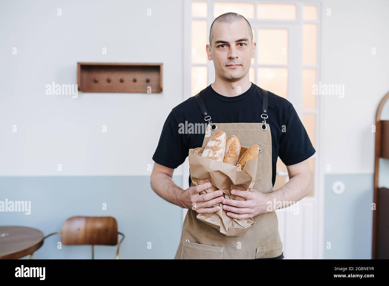 Kräftiger männlicher Bäcker mit Papiertüte und frischen Baguettes. Posiert für ein Foto über einer einfachen Innenumgebung. Stockfoto