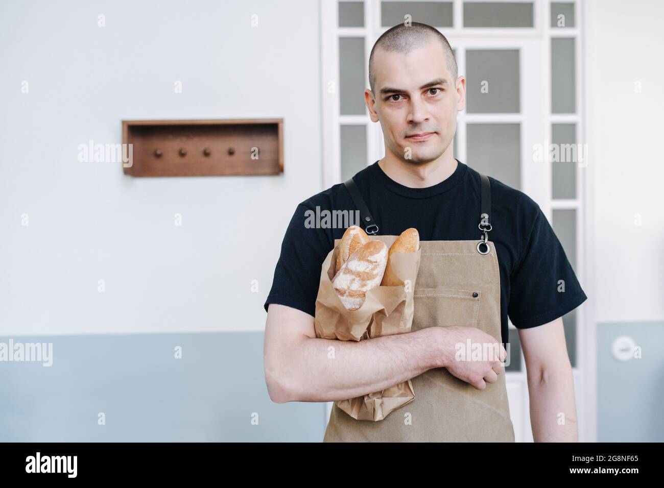 Ernst aussehender männlicher Bäcker, der Papiertüte mit frischen Baguettes hält. Posiert für ein Foto über einer einfachen Innenumgebung. Blick auf die Kamera. Stockfoto