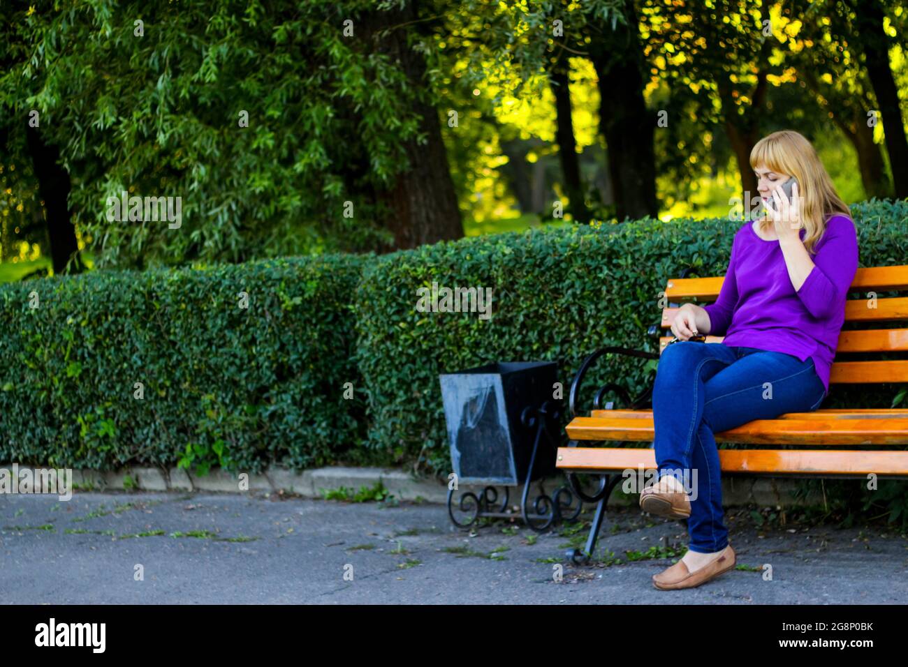 Unschärfe kaukasisch blonde Frau reden, sprechen am Telefon draußen, im Freien. 40-jährige Frau in lila Bluse im Park auf der Bank. Erwachsene weibliche USI Stockfoto
