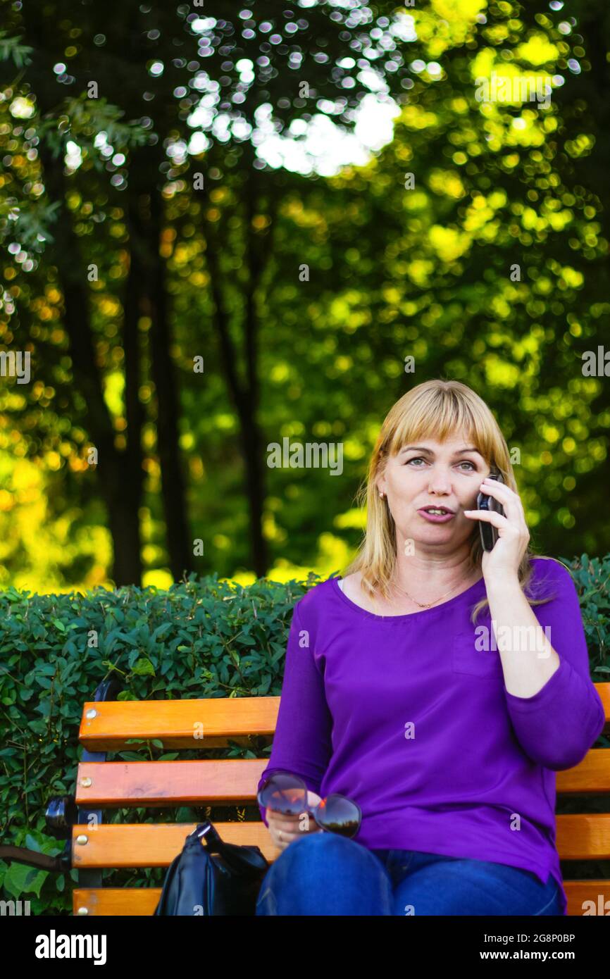 Unschärfe kaukasische Emotion blonde Frau reden, sprechen am Telefon draußen, draußen. 40-jährige Frau in lila Bluse im Park. Erwachsene Person usin Stockfoto