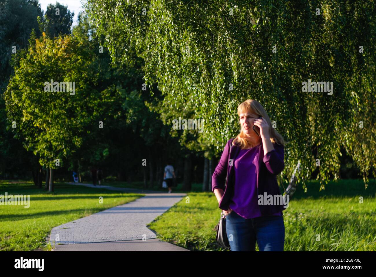 Unschärfe kaukasisch blonde Frau sprechen auf dem Telefon draußen, draußen. 40-jährige Frau in lila Bluse im Park stehend. Erwachsene Frauen, die Telephon benutzen Stockfoto