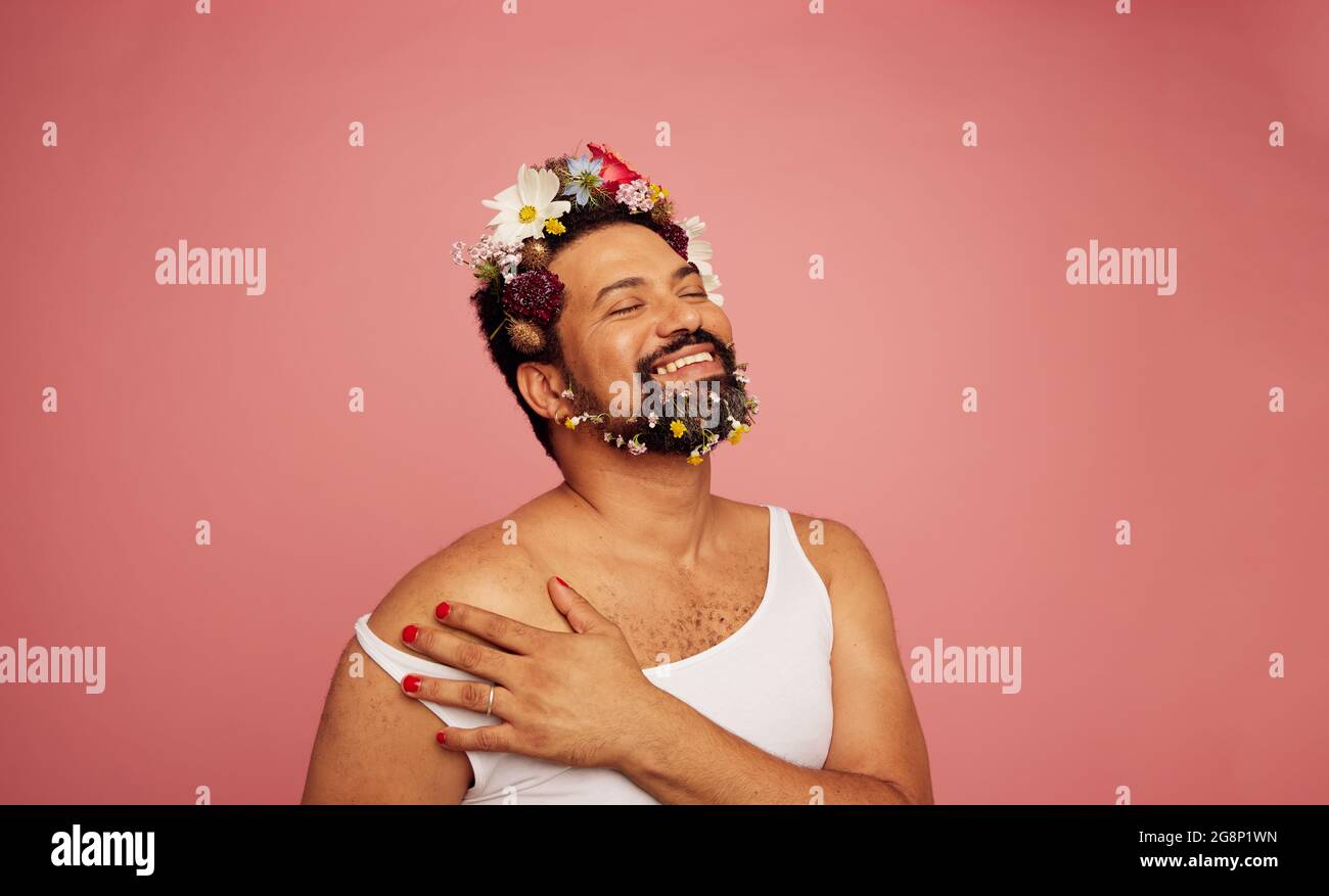 Lächelnde schwule Person auf rosa Hintergrund. Glücklicher bärtiger Mann mit Tanktop und Blumen auf Kopf und Bart. Stockfoto