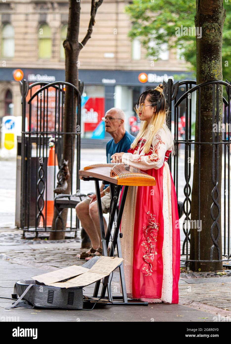 Dundee, Tayside, Schottland, Großbritannien. Juli 2021. UK Wetter: Ein heller, feuchter Tag in Nordostschottland mit Temperaturen von bis zu 21 Grad Eine junge südostasiatische Frau in traditionellem Kleid nimmt den Tag mit, indem sie klassische chinesische Musik mit ihrer Zither spielt, einer Figur, die ein Saiteninstrument im Stadtzentrum von Dundee zupft. Kredit: Dundee Photographics/Alamy Live Nachrichten Stockfoto