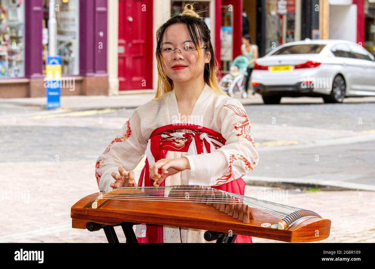 Dundee, Tayside, Schottland, Großbritannien. Juli 2021. UK Wetter: Ein heller, feuchter Tag in Nordostschottland mit Temperaturen von bis zu 21 Grad Eine junge südostasiatische Frau in traditionellem Kleid nimmt den Tag mit, indem sie klassische chinesische Musik mit ihrer Zither spielt, einer Figur, die ein Saiteninstrument im Stadtzentrum von Dundee zupft. Kredit: Dundee Photographics/Alamy Live Nachrichten Stockfoto