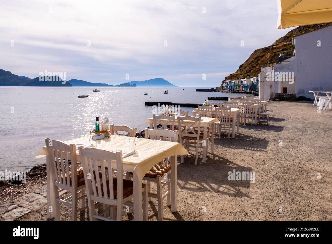 Weiße Esstische an der Küste mit beeindruckender Aussicht aufs Meer und Klima-Fischerdorf im Hintergrund auf der Insel Milos, Griechenland. Stockfoto