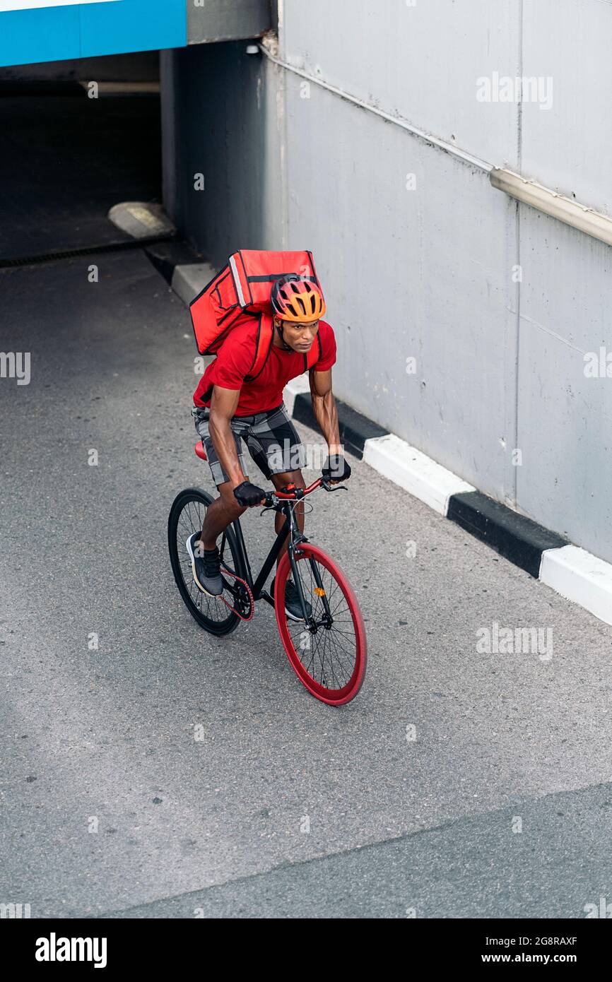 Afrikanischer Lieferer, der ein Paket mit einem Schutzhelm trägt und mit seinem Fahrrad die Stadt überquert. Stockfoto