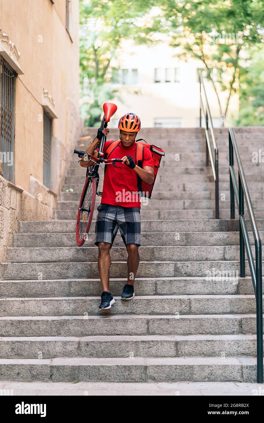 Afrikanischer Lieferer mit Schutzhelm, der sein Fahrrad in der Stadt in der Art und Weise für die Lieferung eines Pakets trägt. Stockfoto