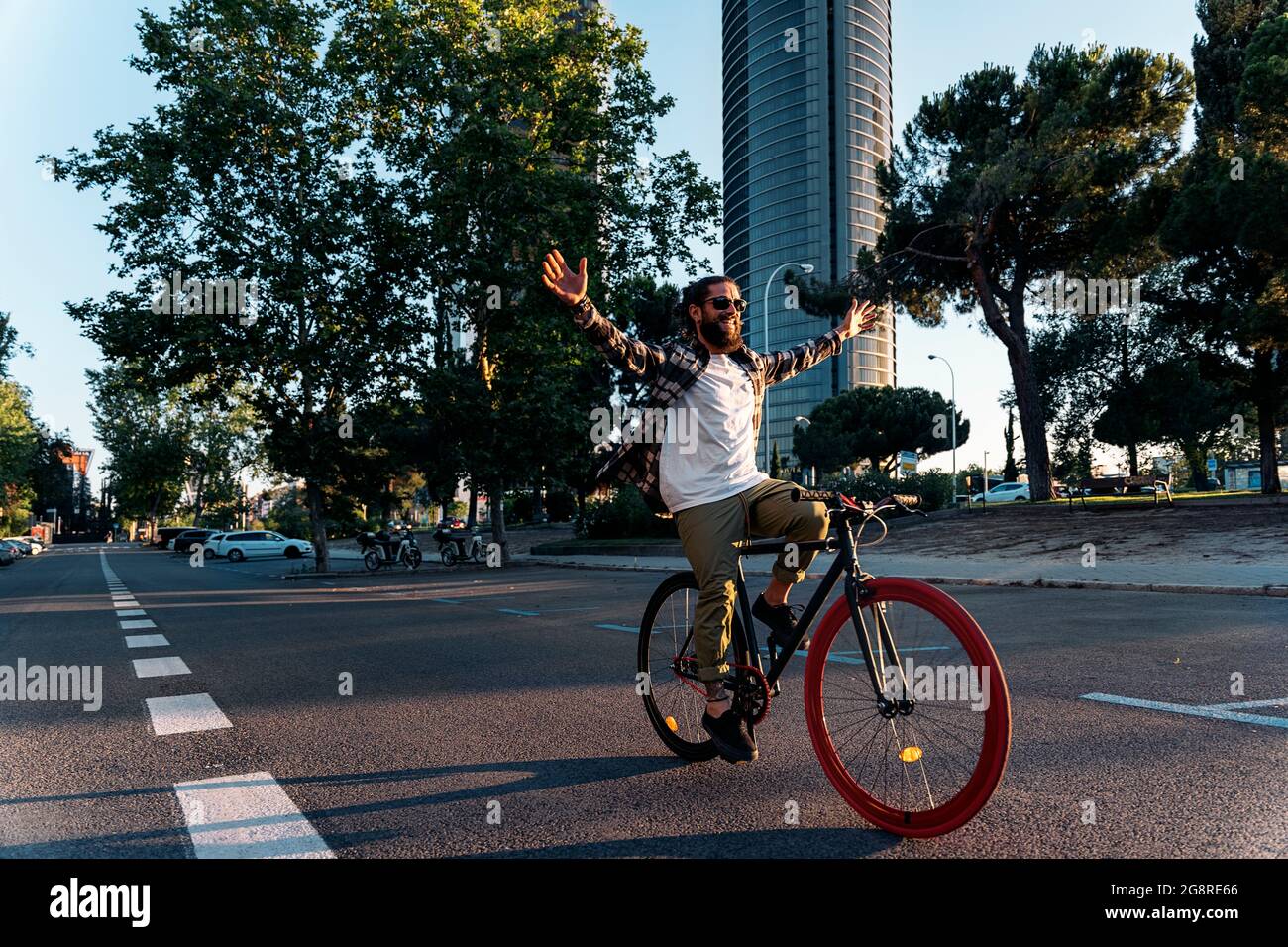 Fröhlicher Hipster-Mann mit großem Bart, der in der Stadt auf dem Fahrrad fährt und die Arme hebt. Stockfoto