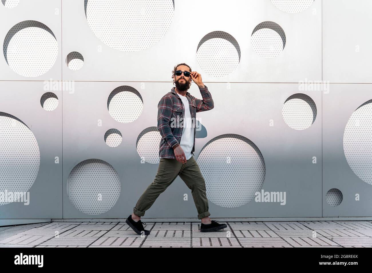 Junger Mann mit langem Bart und Sonnenbrille, der auf der Straße gegen die weiße Wand mit coolem Design läuft. Stockfoto