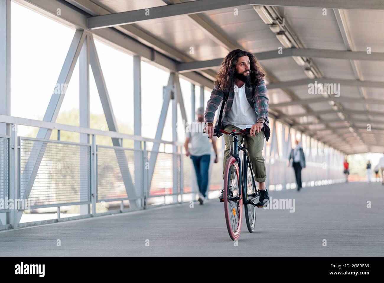 Hübscher Hipster-Mann mit langen Haaren, die zur Seite schauen und mit seinem Fahrrad in der Stadt fahren. Stockfoto