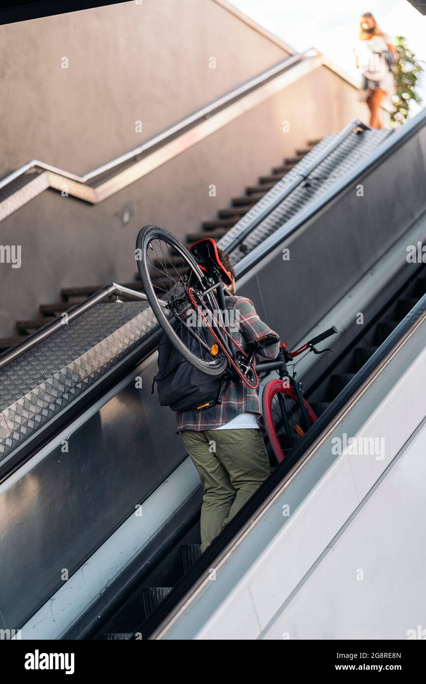 Nicht erkannter Hipster-Mann, der sein Fahrrad trägt, während er die Rolltreppen der U-Bahn hinaufgeht. Stockfoto