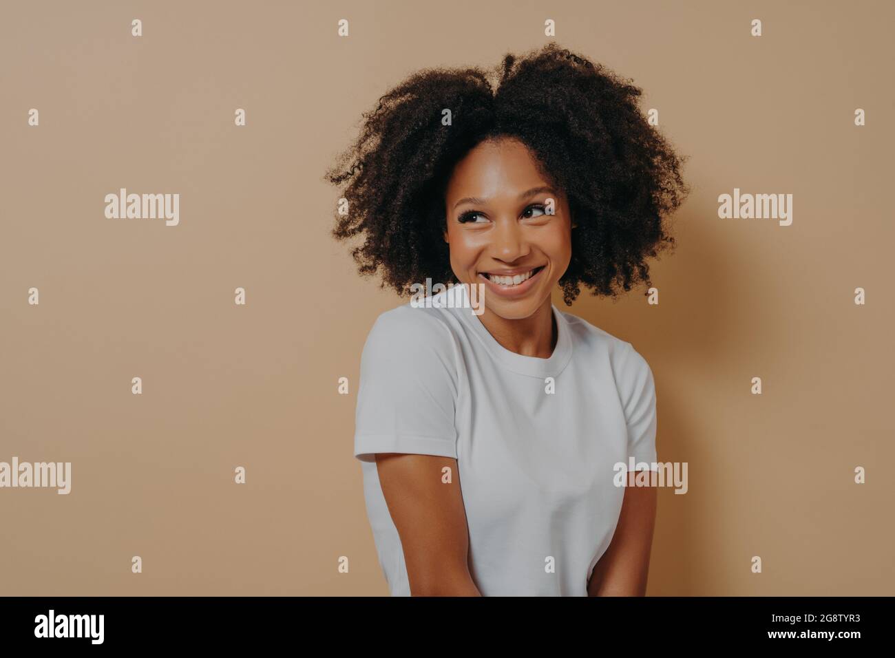 Niedliche afrikanische Frau mit toothy Lächeln Blick beiseite mit Schüchternheit, isoliert auf beigefarbenem Hintergrund Stockfoto