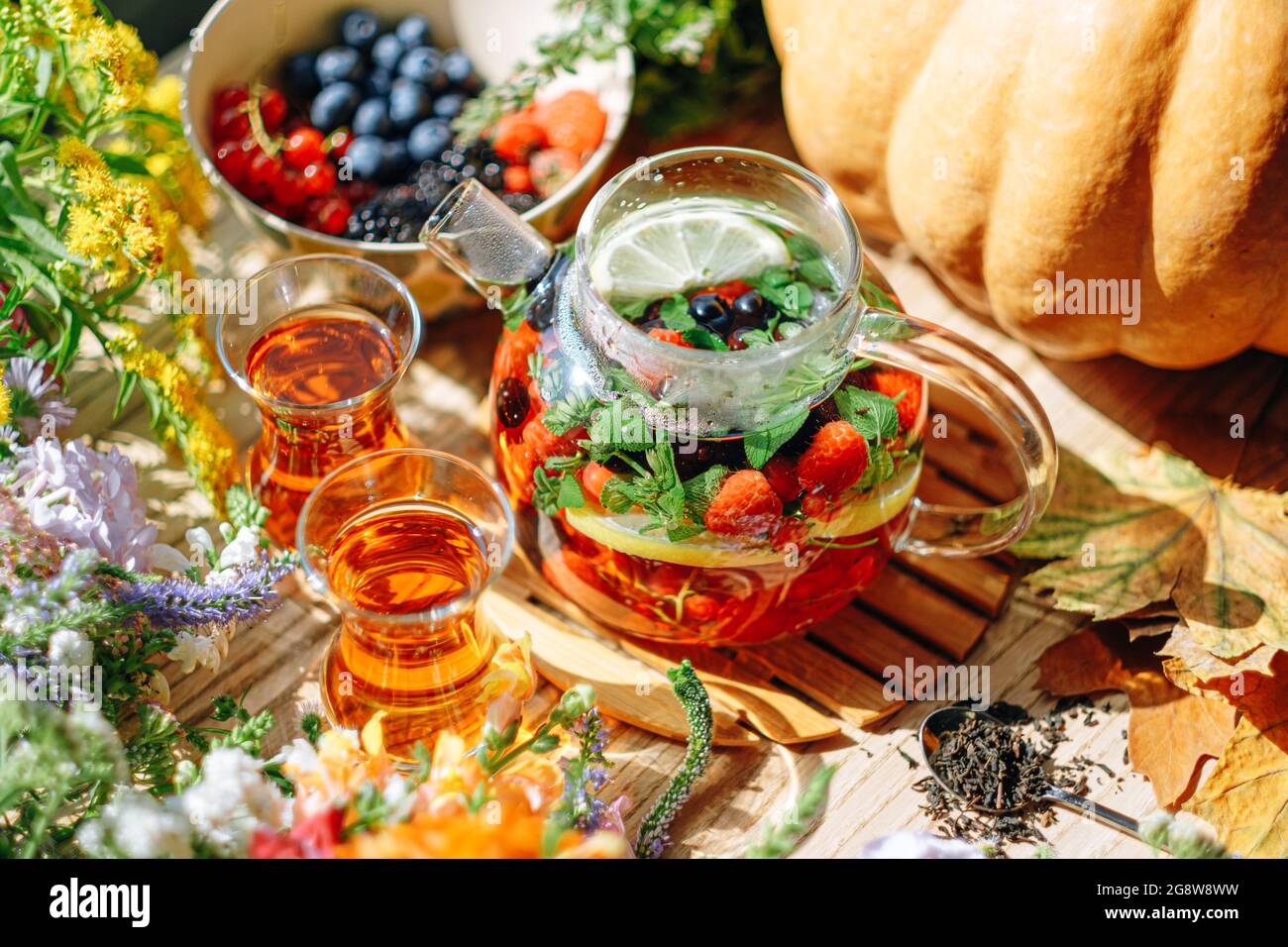 Natürlicher aromatischer Früchtetee in einer transparenten Teekane und in einer Tasse zwischen Beeren und Blumen. Wärmender aromatischer Tee mit einem tiefen Aroma von Beeren und Wildf Stockfoto
