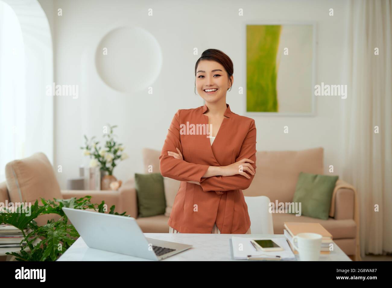 Lächelnde Frau mit verschränkten Armen steht an ihrem Arbeitsplatz im Home Office Stockfoto