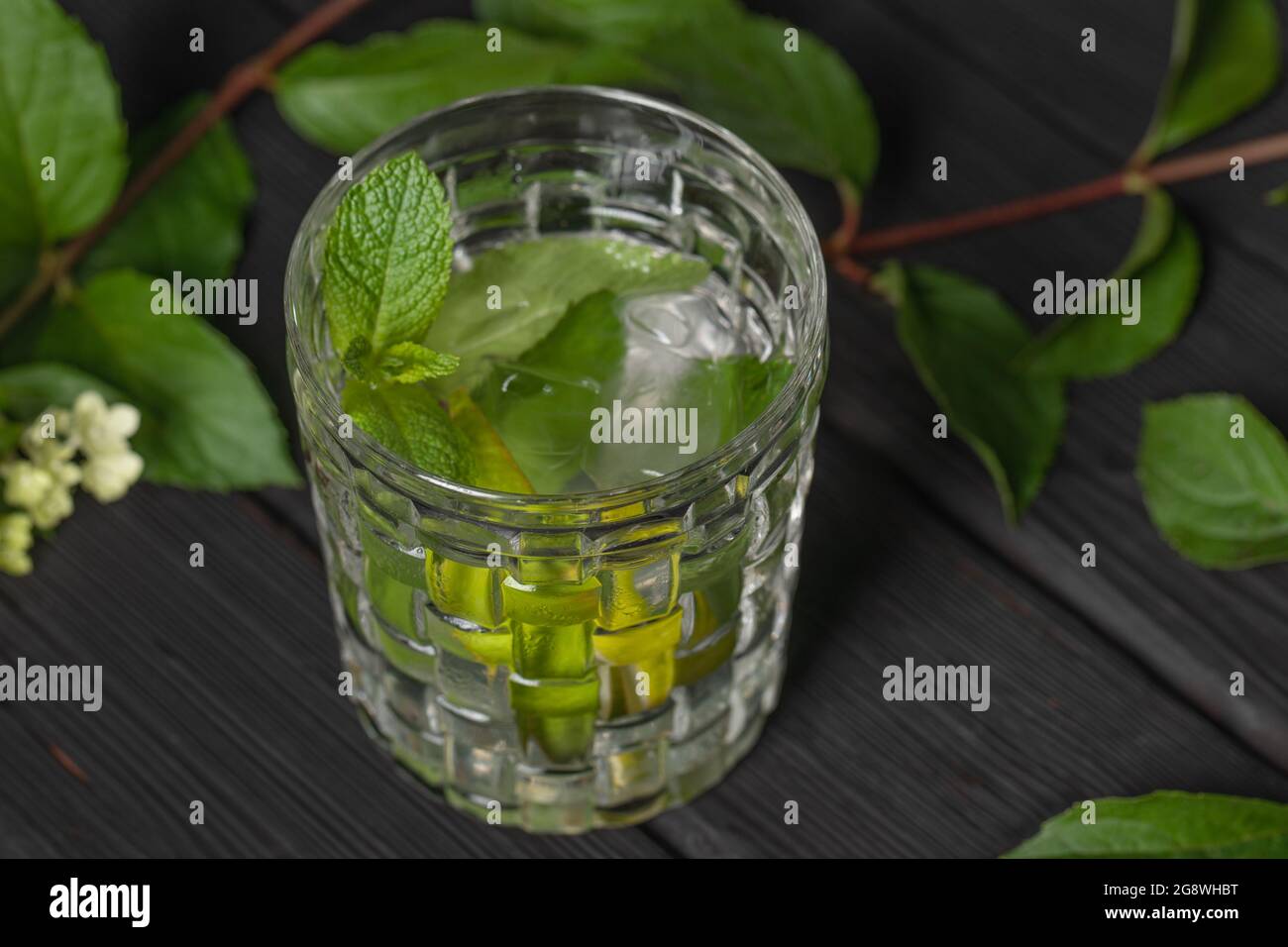 Erfrischender, kalter Mojito-Cocktail mit Limette und Minze im Glas auf verbranntem schwarzem Holzhintergrund, verziert mit Sommerblumen. Stockfoto