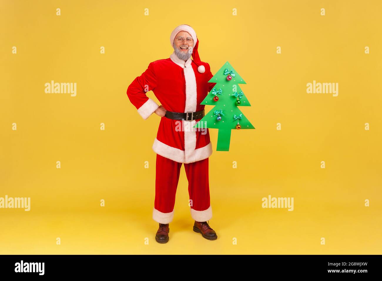 Selbstbewusster erwachsener Mann im weihnachtsmann-Kostüm, der mit der Hand auf der Hüfte steht, einen papiergrünen Weihnachtsbaum hält, blickt lächelnd auf die Kamera. Stehbolzen für den Innenbereich Stockfoto