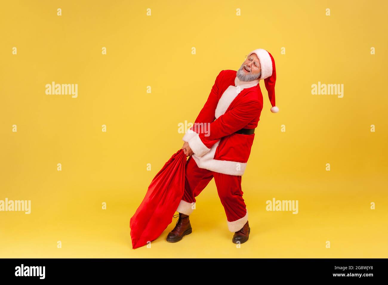 Älterer Mann mit grauem Bart, der ein weihnachtsmann-Kostüm trägt, schleppt seine sehr große und schwere Tasche voller Geschenke, runzelt das Gesicht und versucht, sich zu erheben. Indo Stockfoto
