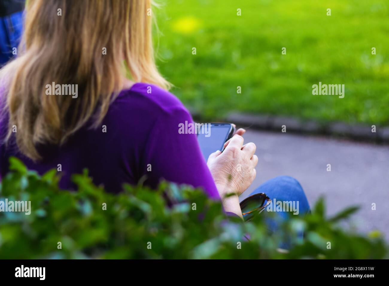 Unschärfe kaukasische blonde Frau sucht auf dem Telefon draußen, im Freien. 40-jährige Frau in lila Bluse im Park mit Smartphone, während sie sitzt Stockfoto