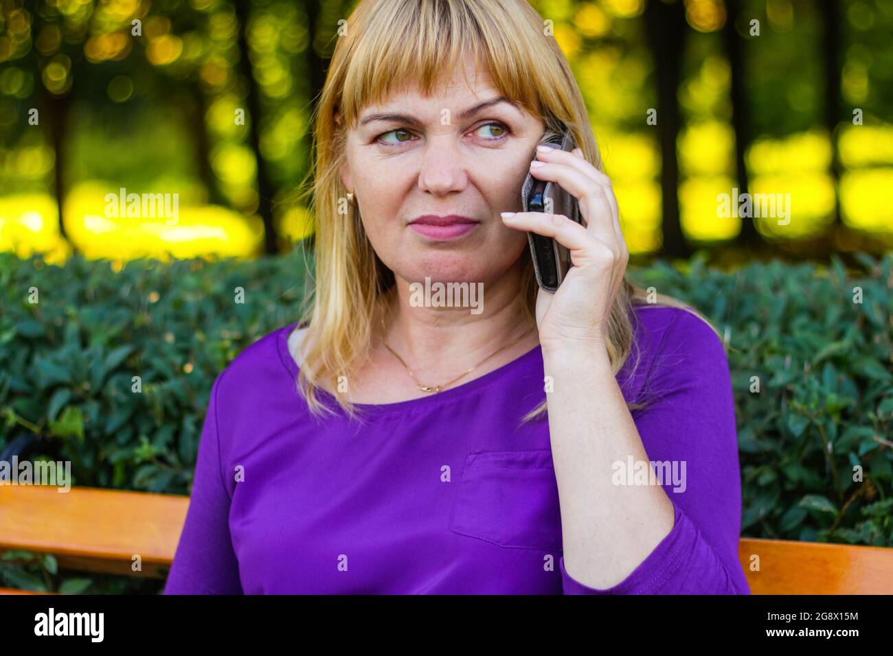 Unschärfe schöne kaukasische blonde Frau sprechen, sprechen auf dem Telefon draußen, im Freien. 45er Jahre alte Frau in lila Bluse im Park. Erwachsene Frauen Stockfoto