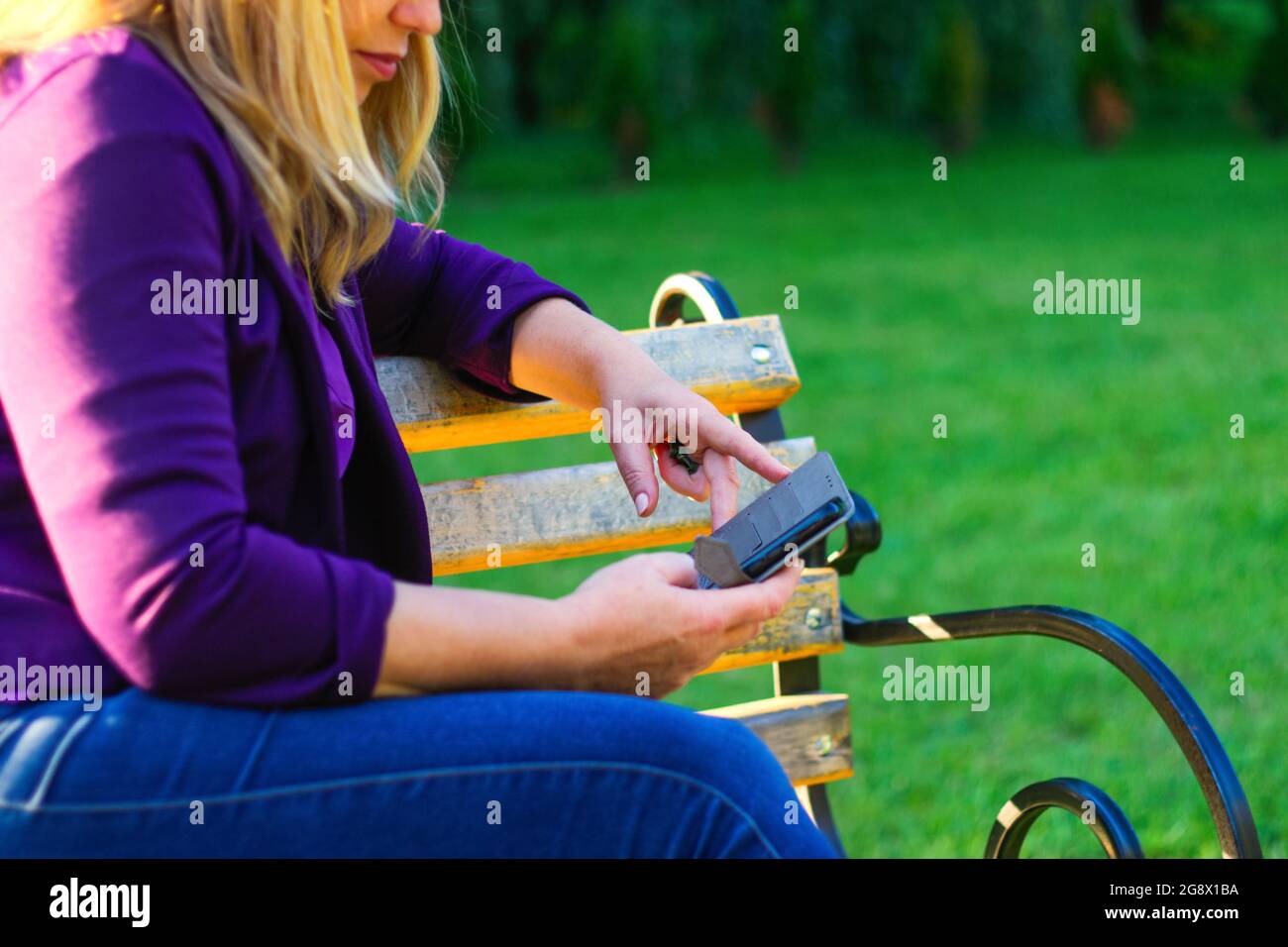 Unschärfe-Nahaufnahme weibliche Frau, die auf das Telefon draußen, im Freien. Frau in lila Bluse mit Smartphone, während sie auf einer Bank im Park sitzt Stockfoto