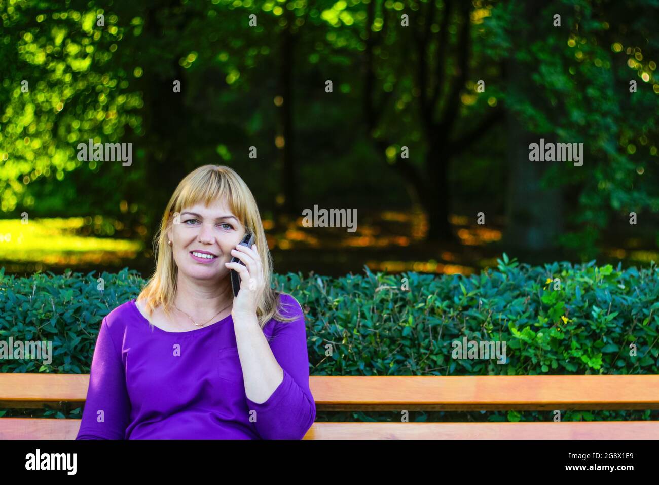 Unschärfe kaukasisch blonde lächelnde Frau sprechen, sprechen auf dem Telefon draußen, draußen. 40-jährige Frau in lila Bluse im Park. Erwachsene, weiblich Stockfoto
