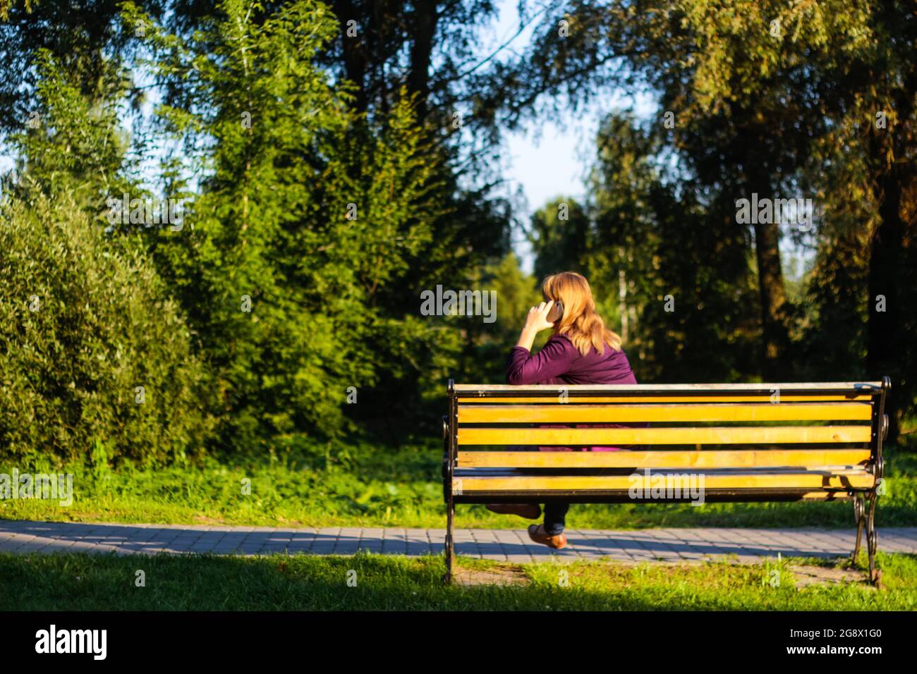 Unschärfe kaukasisch blonde Frau reden, sprechen am Telefon draußen, im Freien. 40-jährige Frau in lila Bluse im Park auf der Bank. Erwachsene Frauen Stockfoto