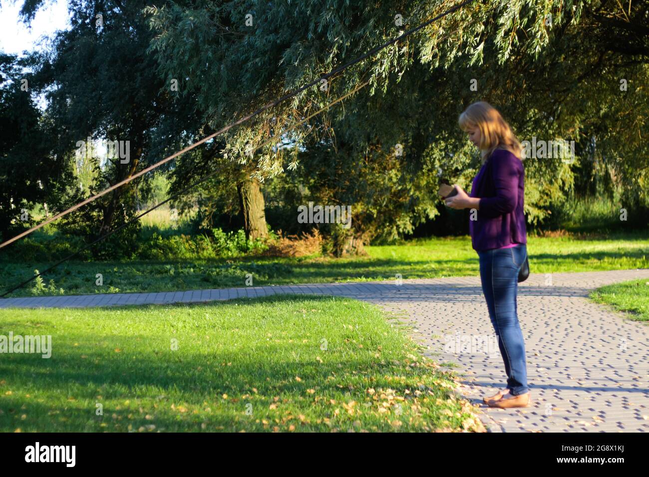 Unschärfe kaukasische blonde Frau sucht auf dem Telefon draußen, im Freien. 40-jährige Frau in lila Bluse im Park stehend. Erwachsene Frauen verwenden Stockfoto