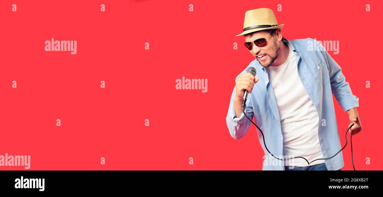 Ein kaukasischer Mann mit Hut und Sonnenbrille singt ein Lied ins Mikrofon. Gesangsleistung des Liedes. Werbebanner für Karaoke-Club oder Bar. Emotionale Sängerin. Studio-Porträt auf rotem Hintergrund. Stockfoto