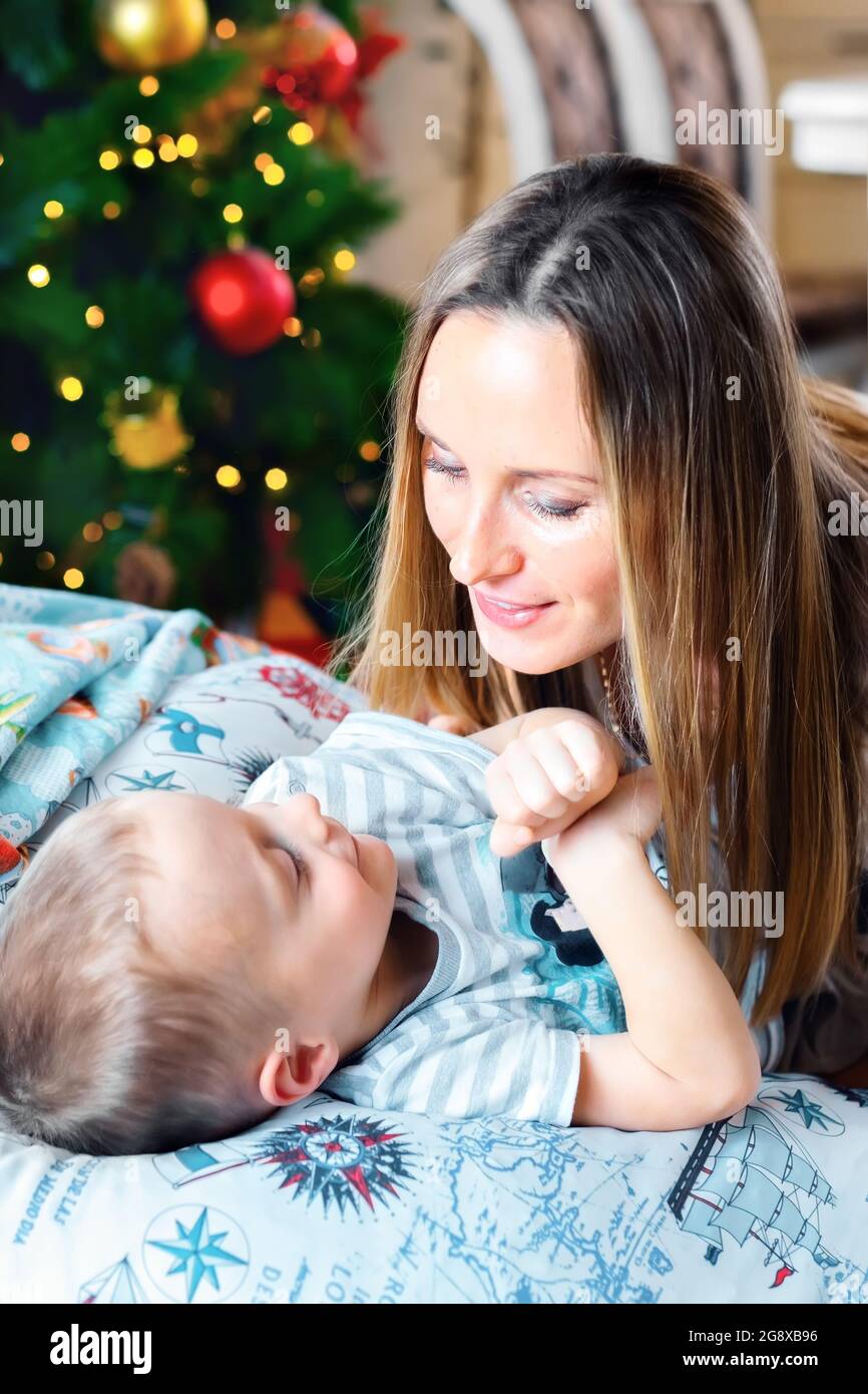 Eine glückliche Mutter mit lockeren Haaren umarmt ihren jungen Sohn vor dem Hintergrund eines Weihnachtsbaums. Warten auf den Urlaub. Liebe und Fürsorge. Aufrichtige Gefühle. Stockfoto