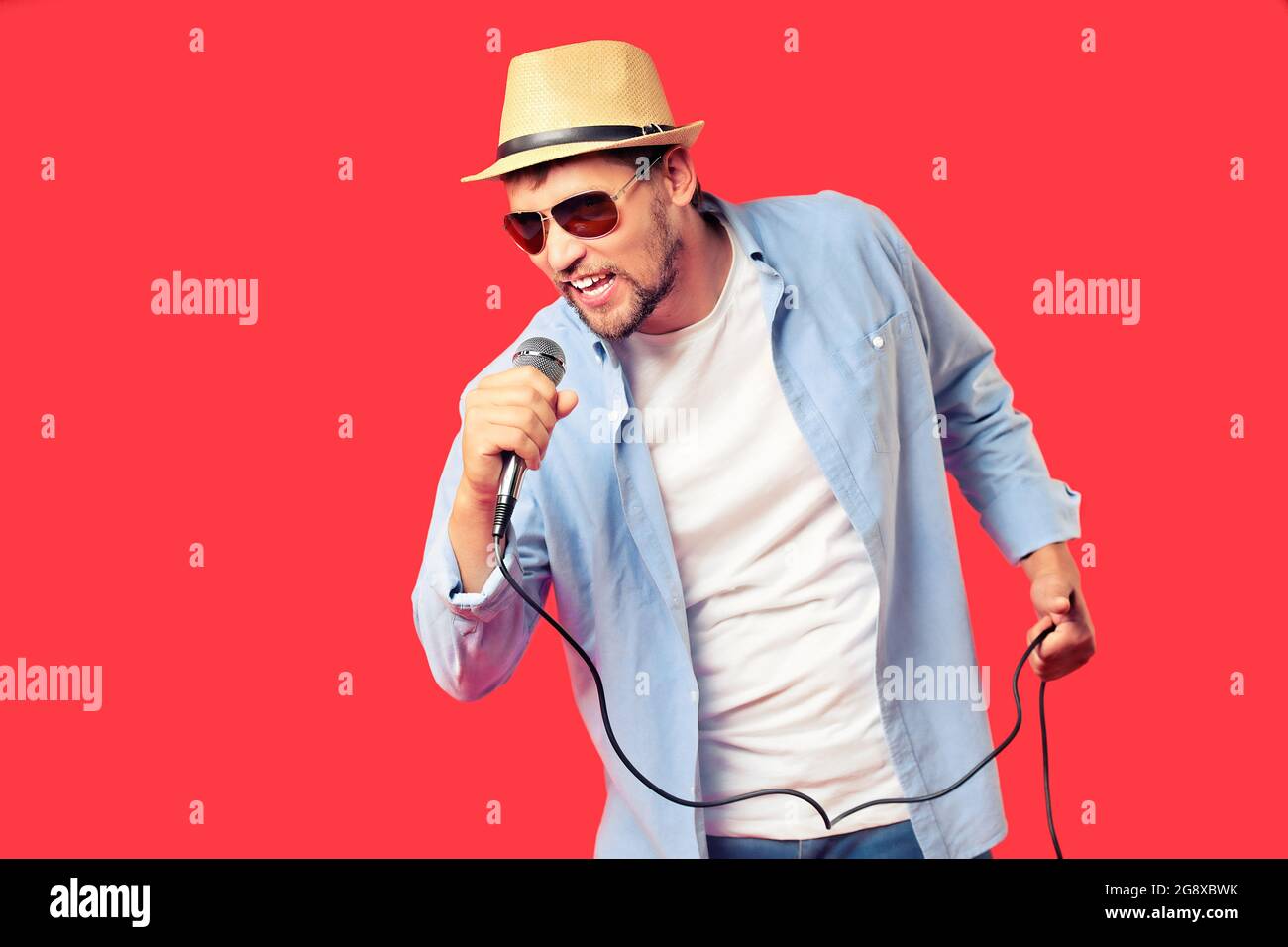 Ein weißer Mann mit Hut und Sonnenbrille singt ein Lied in ein Mikrofon. Gesangsleistung des Liedes. Hintergrund für die Werbung für einen Karaoke-Club oder eine Bar. Eine emotionale Sängerin. Studio-Porträt auf rotem Hintergrund. Stockfoto