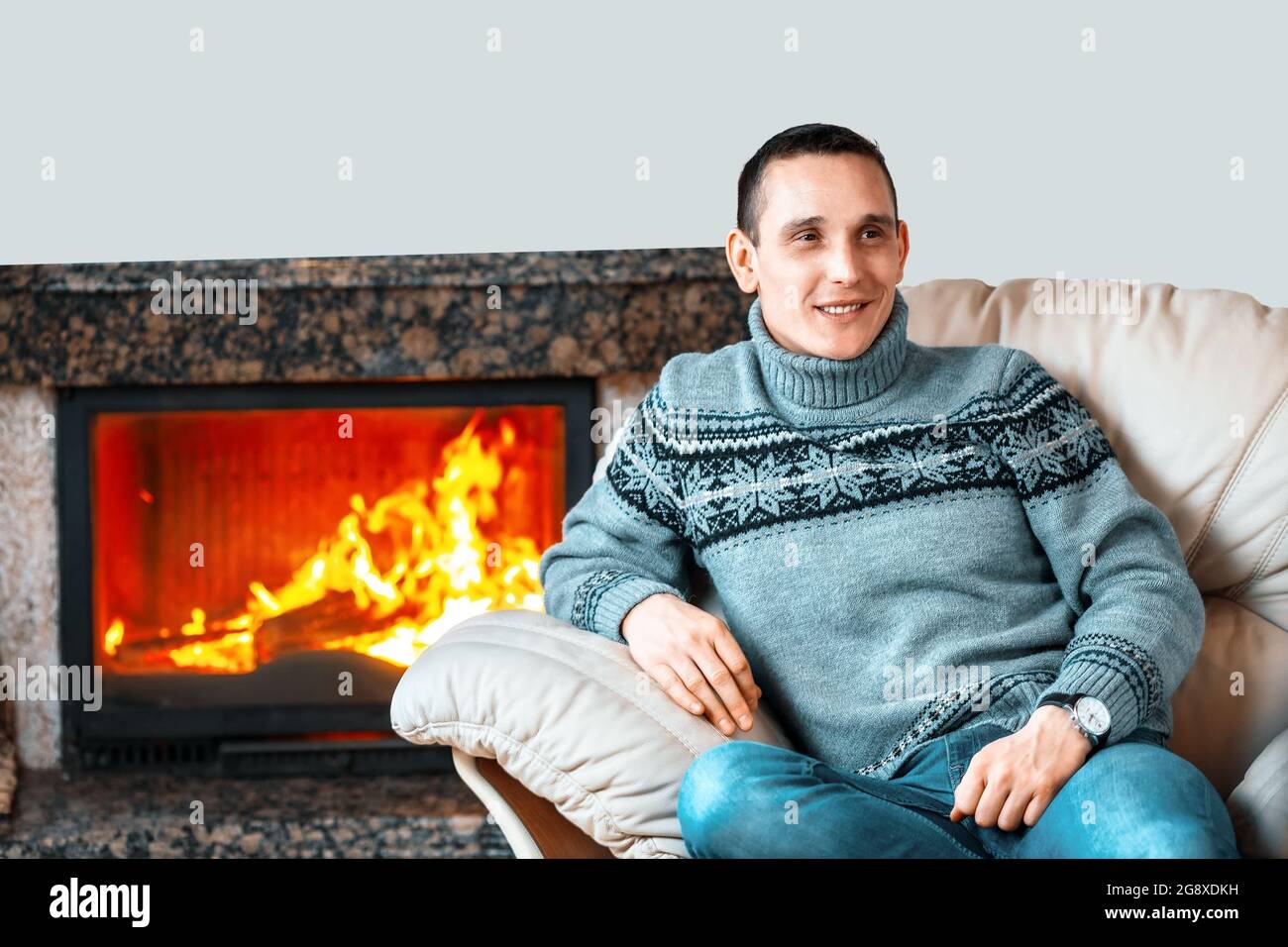 Ein junger Mann in einem Strickpullover sitzt auf einem Lederstuhl vor dem Hintergrund eines Kamins mit Feuer und lächelt. Wohnkomfort und Wärme aus einem luxuriösen Kamin. Copy space Stockfoto