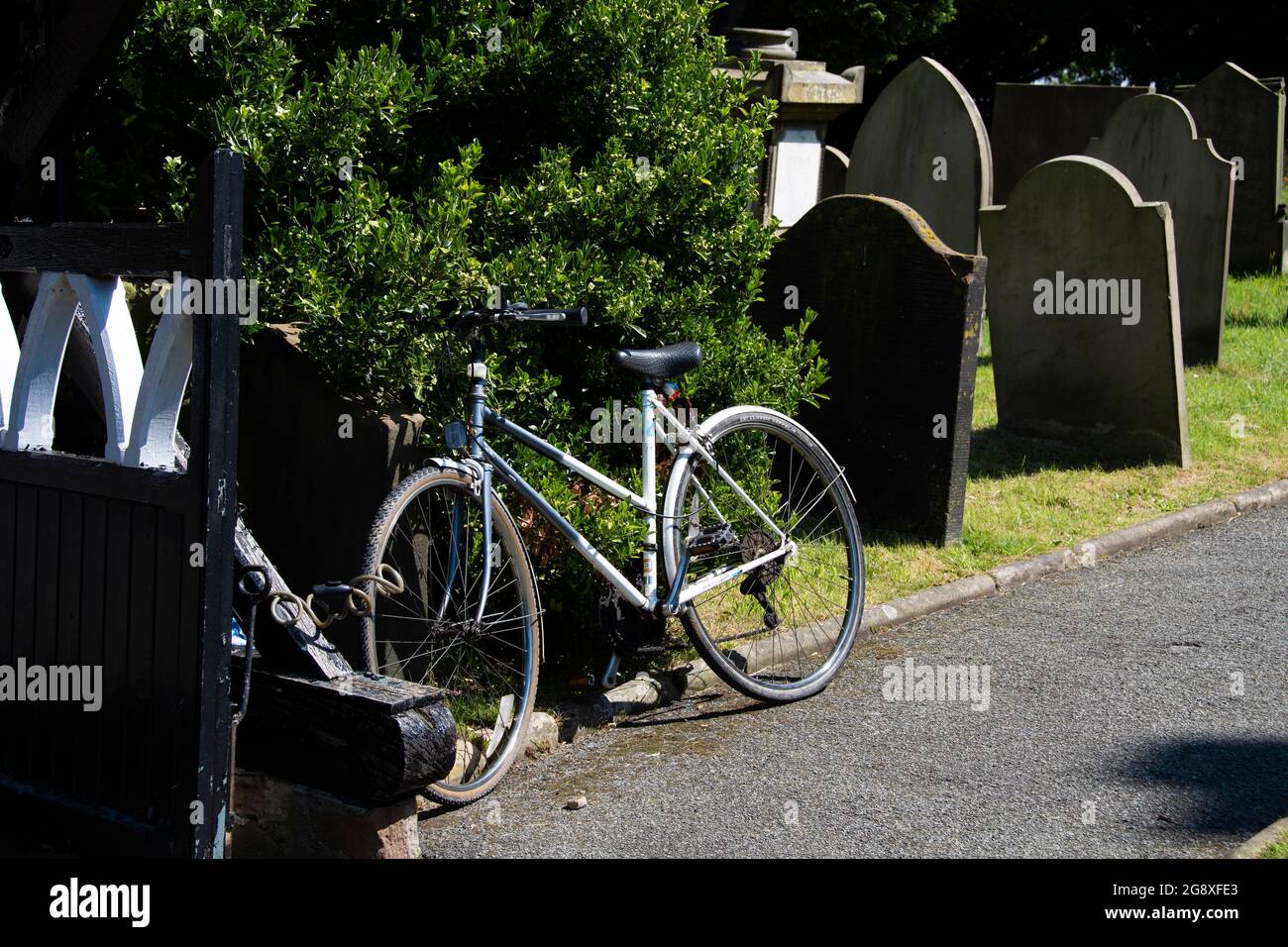 Fahrrad, das auf einem Busch auf einem Friedhof gelehnt ist und am Friedhofstor befestigt ist. Stockfoto