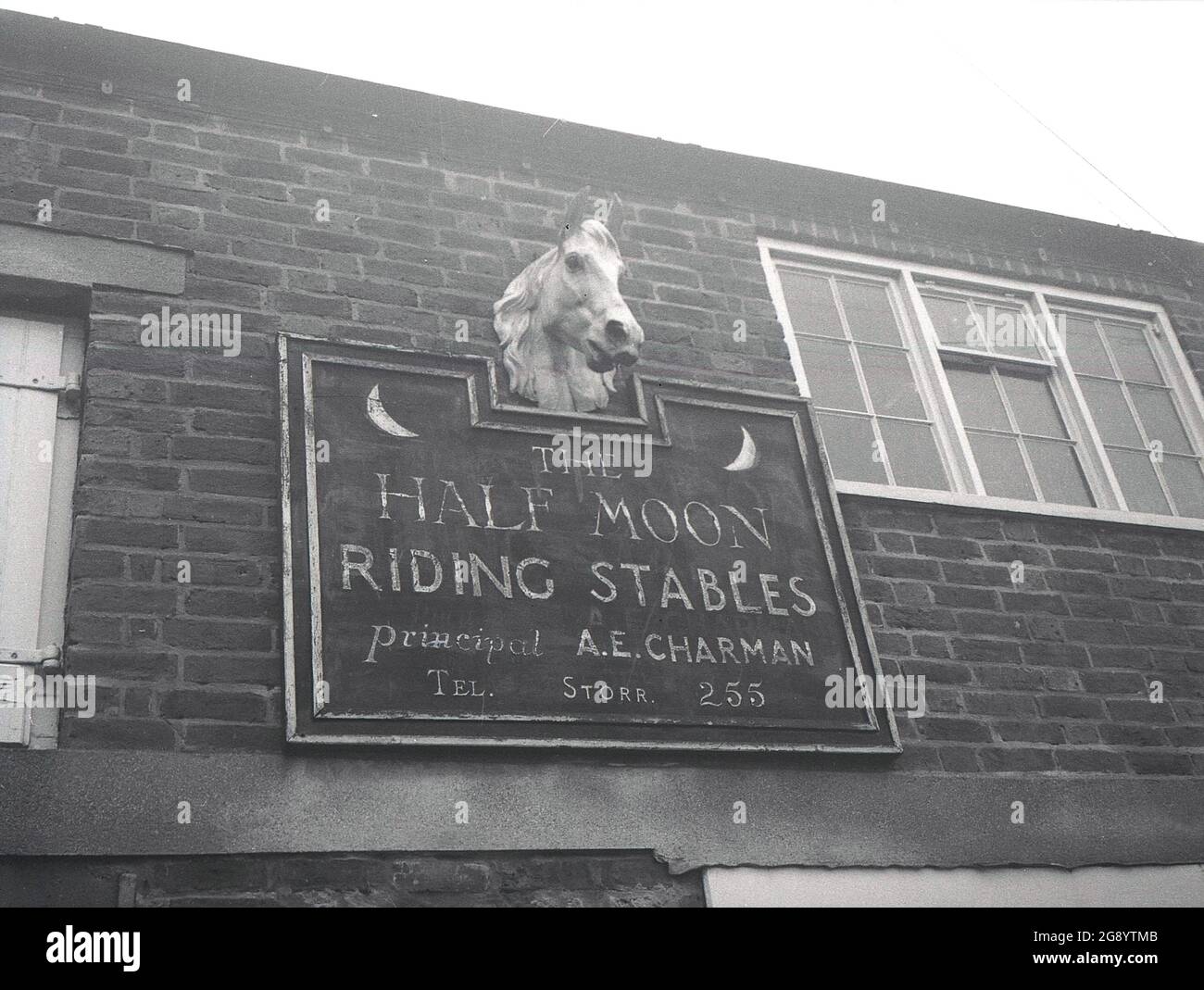 Historisches Zeichen der 1950er Jahre auf der Außenseite für „The Half Moon Riding Stables“ in Storrington, West Sussex, England, Großbritannien. Das Dorf Storrington - oder Storr, wie auf dem Schild kurzgeschlossen - liegt auf der Nordseite des berühmten South Downs Way, einen 100 Meilen langen Spaziergang entlang einer Gegend von außergewöhnlicher natürlicher Schönheit, mit Blick auf den Ärmelkanal. Stockfoto