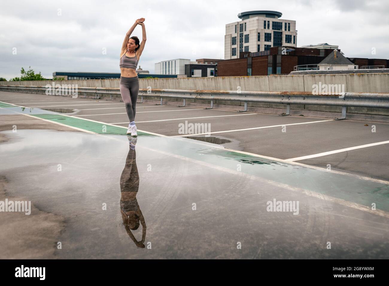 Fit sportliche Frau macht Dehnübungen draußen auf einem städtischen Parkplatz mit Pfütze und Reflexion. Stockfoto