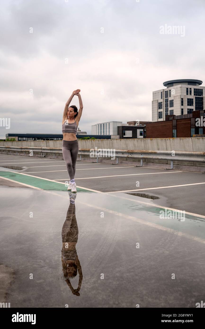 Fit sportliche Frau macht Dehnübungen draußen auf einem städtischen Parkplatz mit Pfütze und Reflexion. Stockfoto