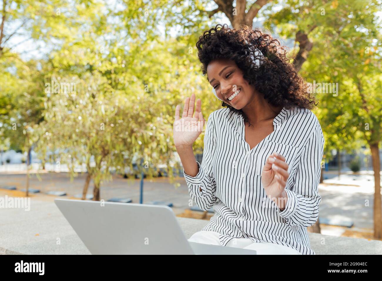 Eine glückliche, lockige Geschäftsfrau winkt, während sie im Büropark einen Videoanruf über den Laptop führt Stockfoto