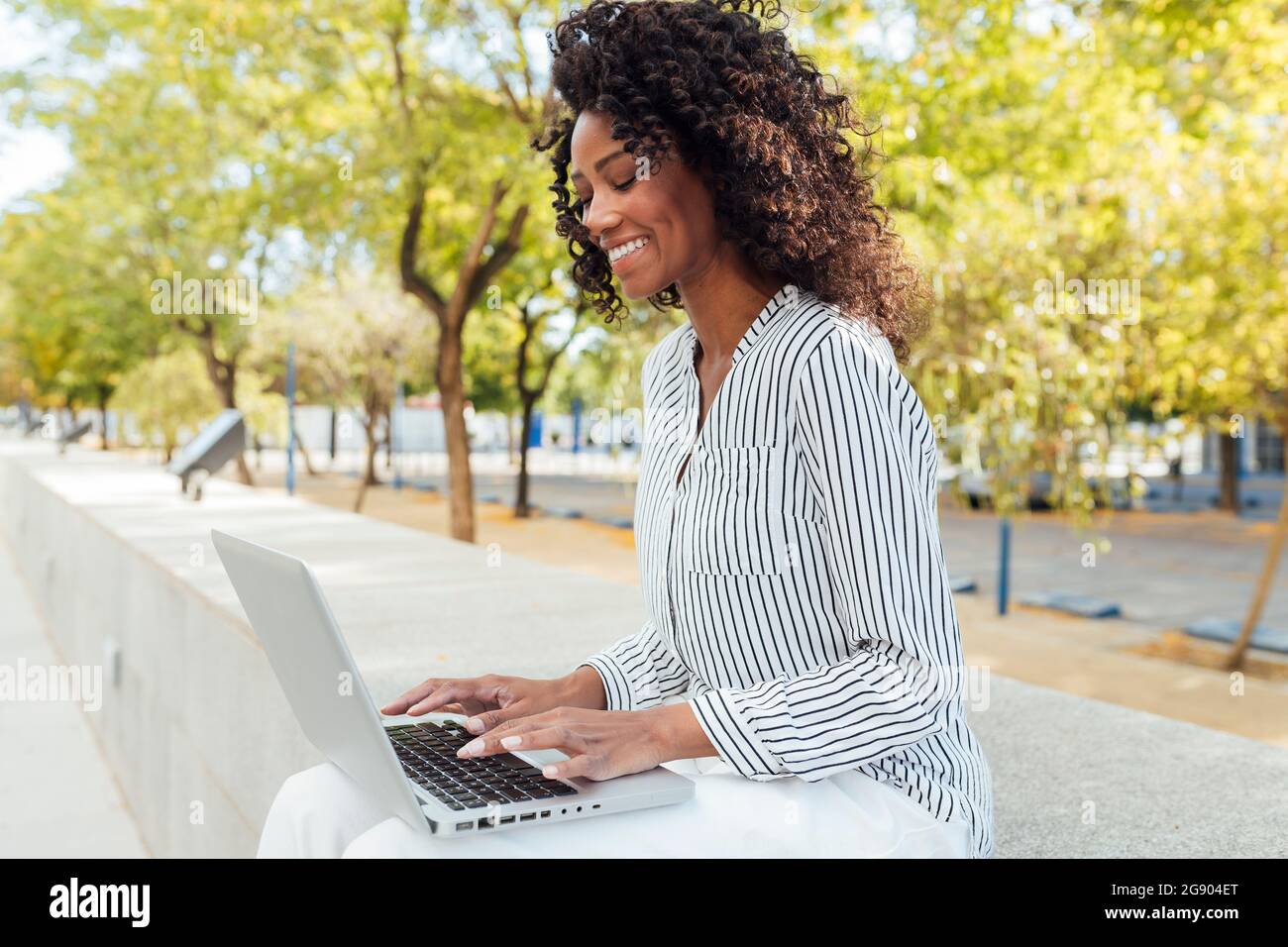 Lächelnde, lockige Geschäftsfrau, die mit einem Laptop am Büropark sitzt und tippt Stockfoto