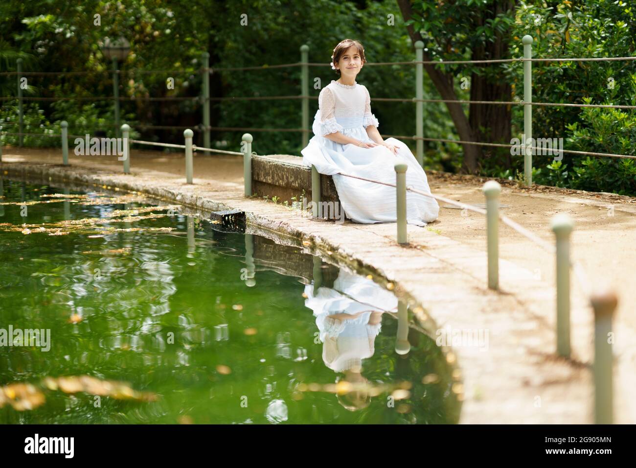 Mädchen in weißem Kleid sitzt auf Felsen am Teich im Garten Stockfoto