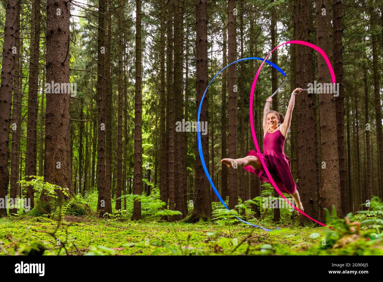 Lächelnde weibliche Turnerin macht rhythmische Gymnastik mit Bändern im Wald Stockfoto