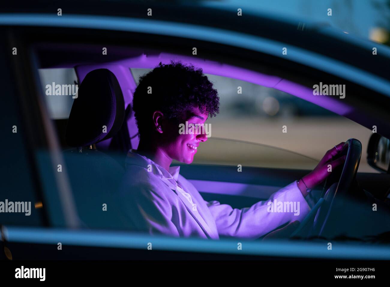 Lächelnde Geschäftsfrau, die nachts in einem beleuchteten, violetten Auto sitzt Stockfoto