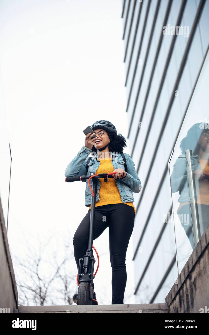Frau mit Elektro-Push-Roller senden Sprachnachricht über das Smartphone durch Gebäude Stockfoto